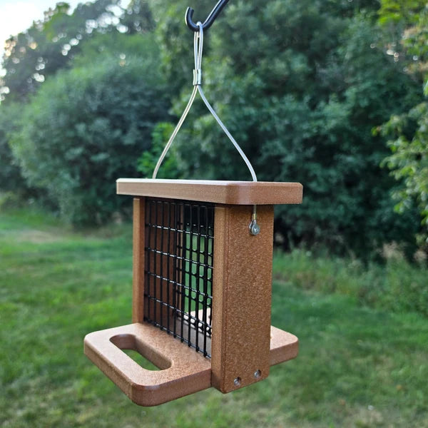 A brown hanging bird feeder with a black wire grid and metal hook, photographed outdoors against a blurred green garden background.
